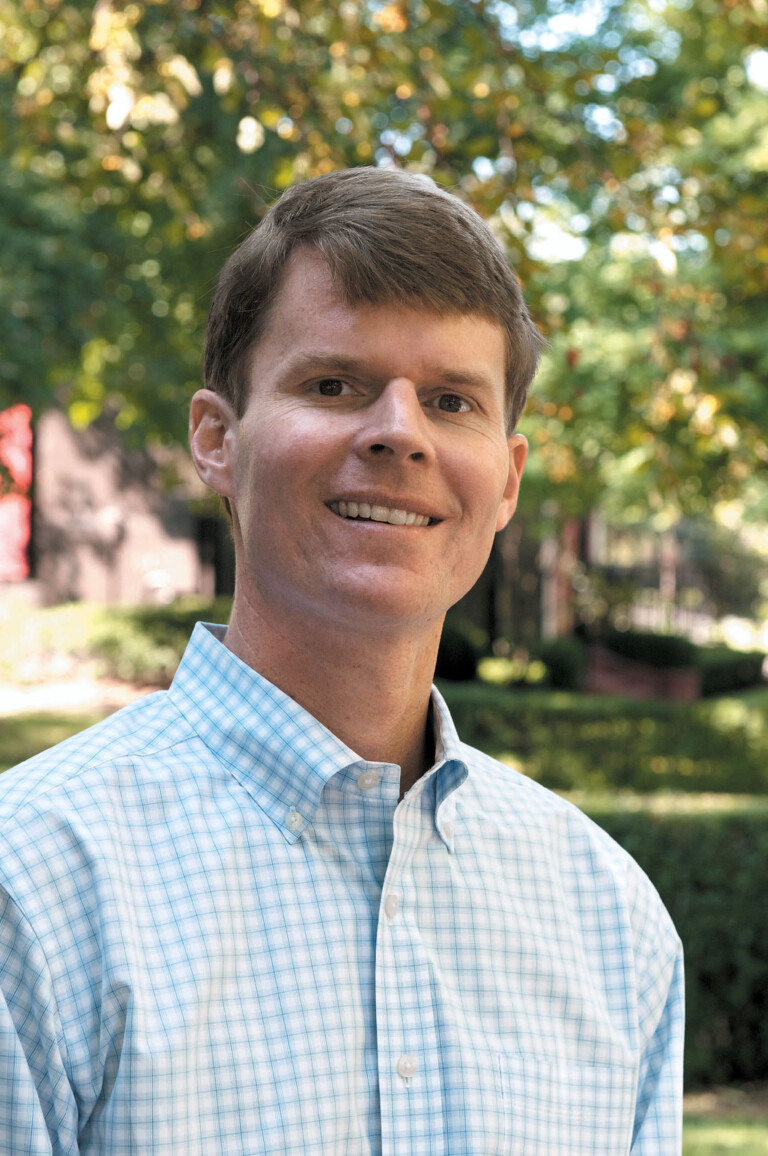 A man with short brown hair smiling, wearing a light blue checkered button-up shirt, standing outdoors with greenery in the background.