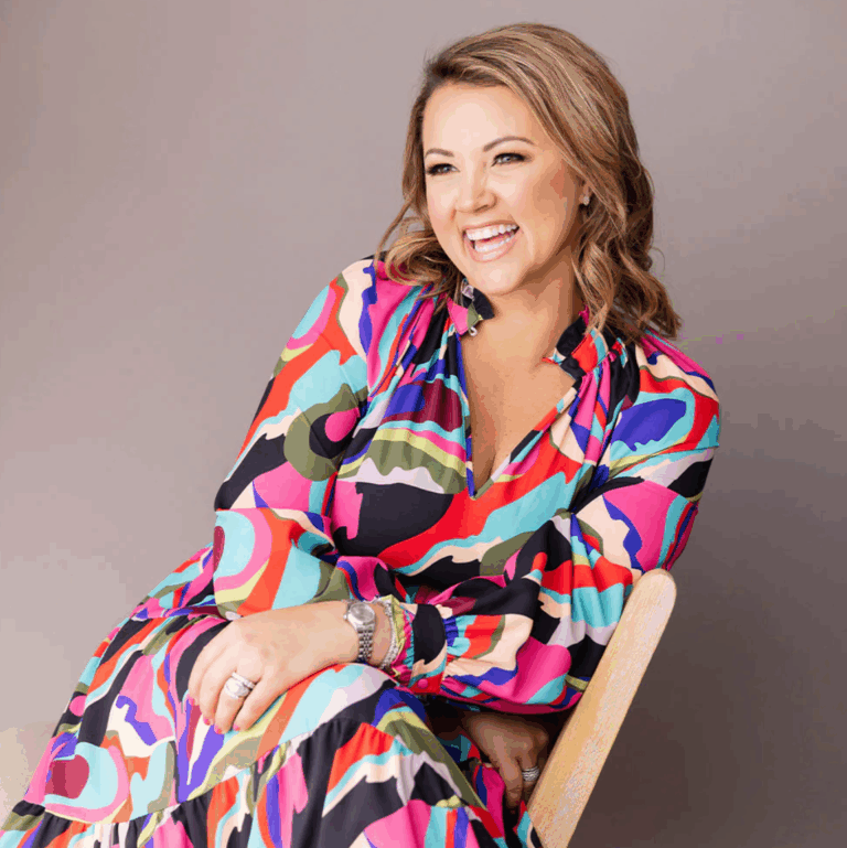 A woman with wavy hair is smiling while sitting on a light-colored chair, wearing a colorful, abstract-patterned dress with shades of pink, green, and black. The background is a soft gray.