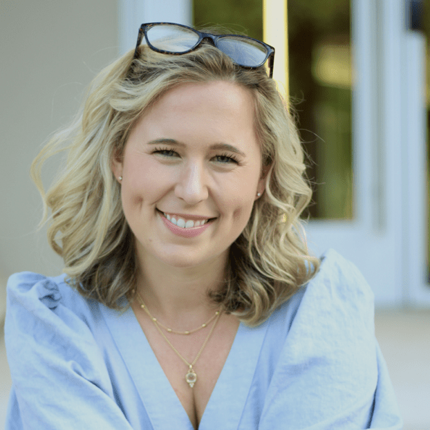 A smiling woman with wavy blonde hair wears a light blue top and three layered necklaces, sitting outside with her arms crossed, and glasses resting on her head, with a blurred entrance in the background.