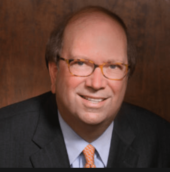 A smiling man with glasses and light brown hair, wearing a black suit and a white collared shirt with an orange tie, posed against a dark wooden background.