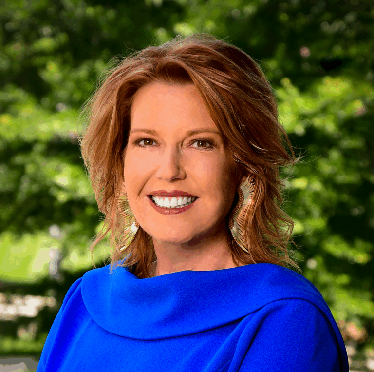 A woman with shoulder-length wavy hair wearing a bright blue blouse and large decorative earrings, smiling warmly against a softly blurred green background of foliage.