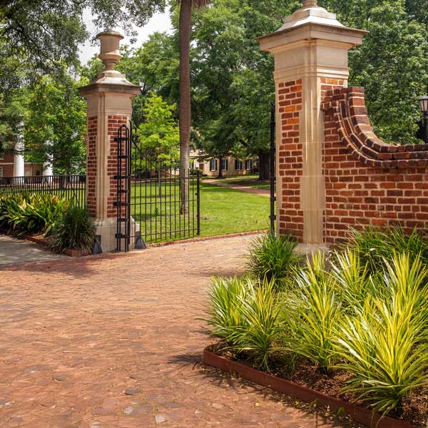 Decorative brick pillars with wrought iron gates lead into a manicured green park area, with palm trees and colorful shrubs framed by a brick pathway.