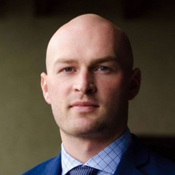 A man with a bald head wearing a blue suit and a checkered shirt, standing against a blurred background.