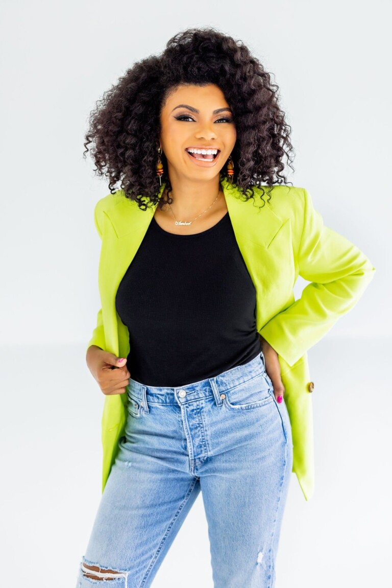 A smiling woman with curly black hair wearing a bright lime green blazer over a black top, paired with light blue distressed jeans, stands against a light gray background.