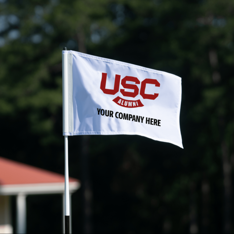 A white flag displaying the text 'USC ALUMNI' in red letters above the phrase 'YOUR COMPANY HERE' in black letters, set against a blurred green background of trees.