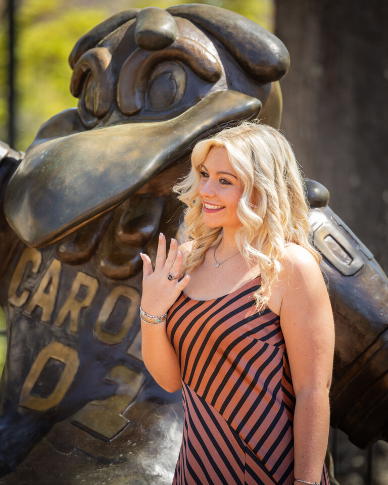 A young woman with wavy blonde hair, wearing a striped dress, stands smiling in front of a bronze statue of a bird mascot, raising one hand to display a ring while posing outdoors on a sunny day.
