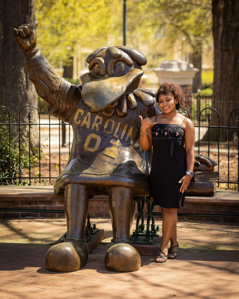 A young woman stands next to a large bronze statue of a bird mascot wearing a jersey labeled 'CAROLINA 02.' The woman, with curly hair, is making a hand gesture while smiling and wearing a black dress. The background features trees and a park-like setting.