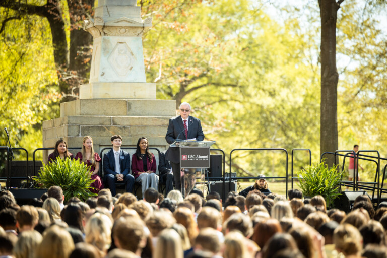 A speaker at a podium addresses an audience seated outdoors, with a monument visible in the background. Four individuals in formal attire sit beside the speaker, and a large audience is gathered in front, with greenery around the scene.