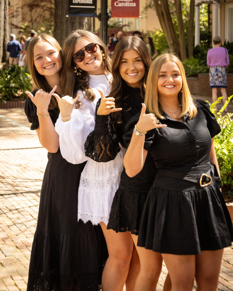 Four young women pose together outdoors, wearing stylish dresses in black and white, with the middle one wearing sunglasses and making a hand gesture. They stand in front of greenery and a brick pathway, conveying a joyful and friendly atmosphere.