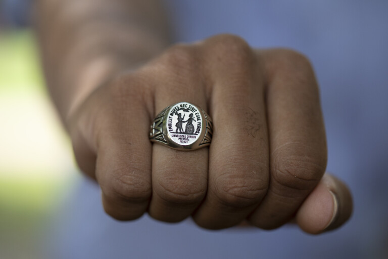 A close-up of a clenched fist wearing a decorative silver ring, featuring an emblem and text that reads 'Universitas Carolina 1801'. The hand is positioned against a blurred background.