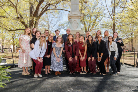 A group of 20 people posing for a photo outdoors near a monument, featuring individuals in various outfits, including formal and casual wear, with trees and buildings in the background under clear blue skies.