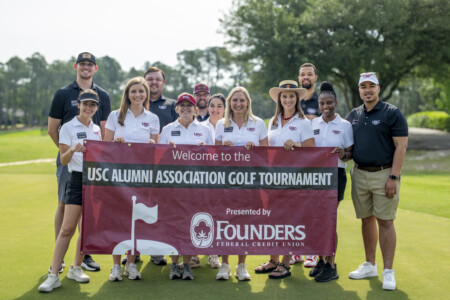 A group of people holding a large banner that reads 'Welcome to the USC Alumni Association Golf Tournament' while posing on a golf course.