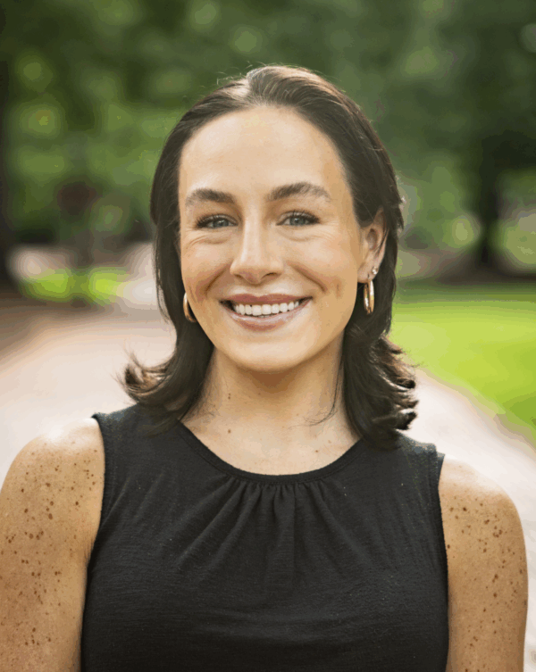 A smiling woman with medium-length brown hair and freckles, wearing a black sleeveless top, stands in a green outdoor setting, with a blurred pathway behind her.