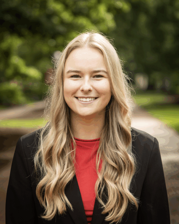 A young woman with long, wavy blonde hair smiles confidently at the camera, wearing a black blazer over a red top, set against a blurred background of green trees and a pathway.