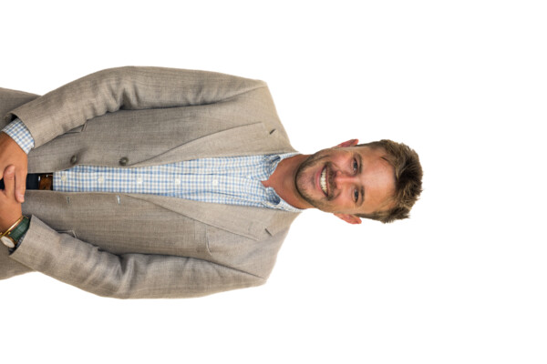 A man with short dark hair and a trimmed beard smiles while standing in front of a plain white background. He wears a light gray blazer over a blue and white checked shirt and is posed with his hands clasped in front of him.