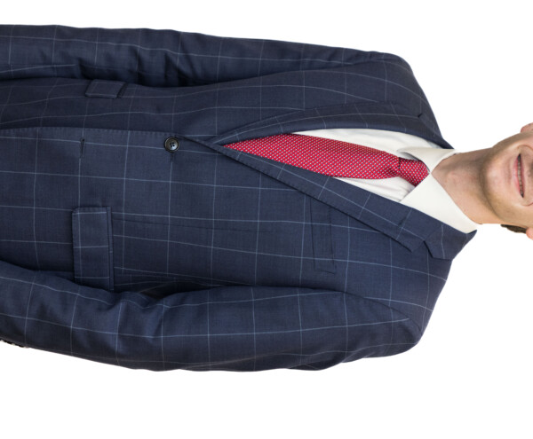A young man in a navy blue checkered suit and a red tie, standing and smiling against a white background.