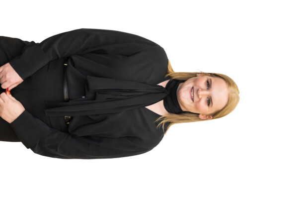 A smiling woman stands confidently in a black outfit featuring a long-sleeve blouse with a scarf detail and black pants, with her hands clasped in front of her.
