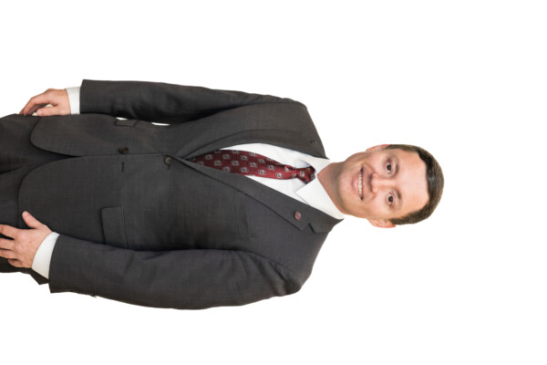 A man in a gray suit and a maroon tie with a pattern stands smiling against a white background. He has short brown hair and is wearing a white dress shirt.