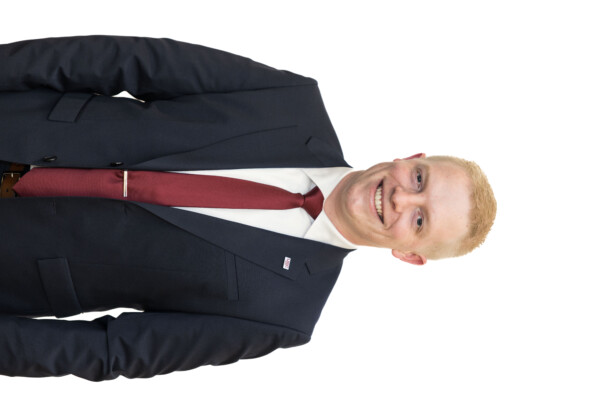 A smiling man in a dark suit and a maroon tie, standing against a white background. He has short blonde hair and a pin on his suit jacket.