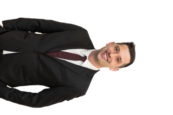 A man in a formal black suit with a maroon tie smiles at the camera, standing against a white background. He has short hair and appears friendly and professional.
