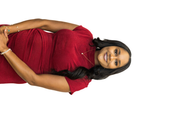 A woman smiling while wearing a red dress, standing against a white background. She has long, wavy hair and is wearing subtle jewelry, including a necklace and bracelets.
