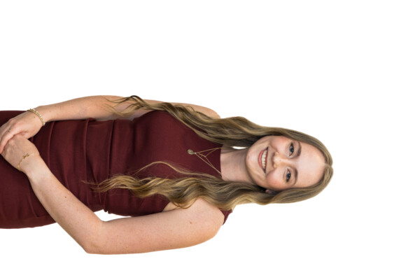 A young woman with long, wavy hair wearing a burgundy sleeveless dress, standing against a white background and smiling, with her hands clasped in front of her.