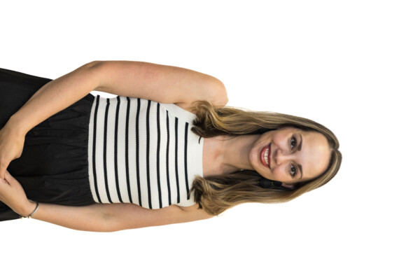 A woman with long, wavy hair stands smiling, wearing a sleeveless striped top with black and white stripes and a black skirt. She has a bracelet on her wrist and is positioned against a plain white background.