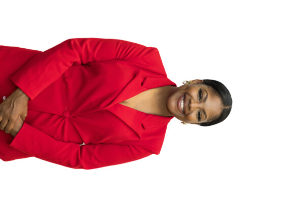 A smiling woman in a bright red blazer with a notched collar stands against a plain white background, with her hands clasped in front of her.