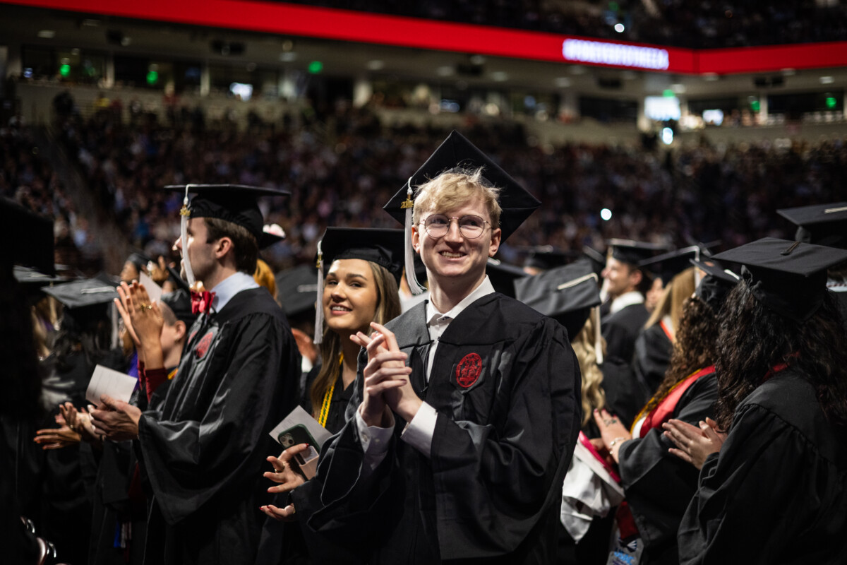 A group of graduates in black caps and gowns applaud during a commencement ceremony, with a focus on a smiling male graduate with glasses in the foreground.