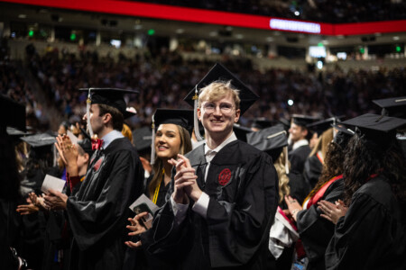 A group of graduates in black caps and gowns applaud during a commencement ceremony, with a focus on a smiling male graduate with glasses in the foreground.