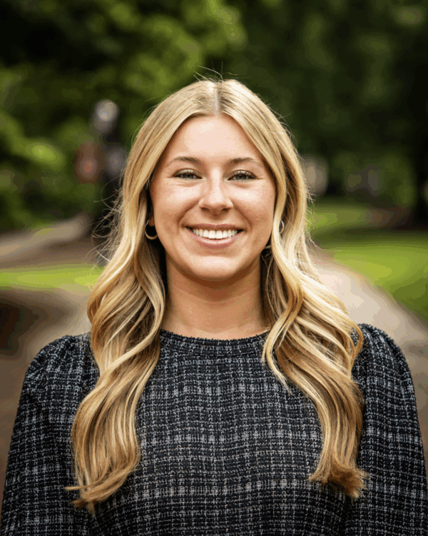 A woman smiling at the camera, with long blonde hair and wearing a black patterned sweater. She stands outdoors with a blurred green background of trees and a pathway.