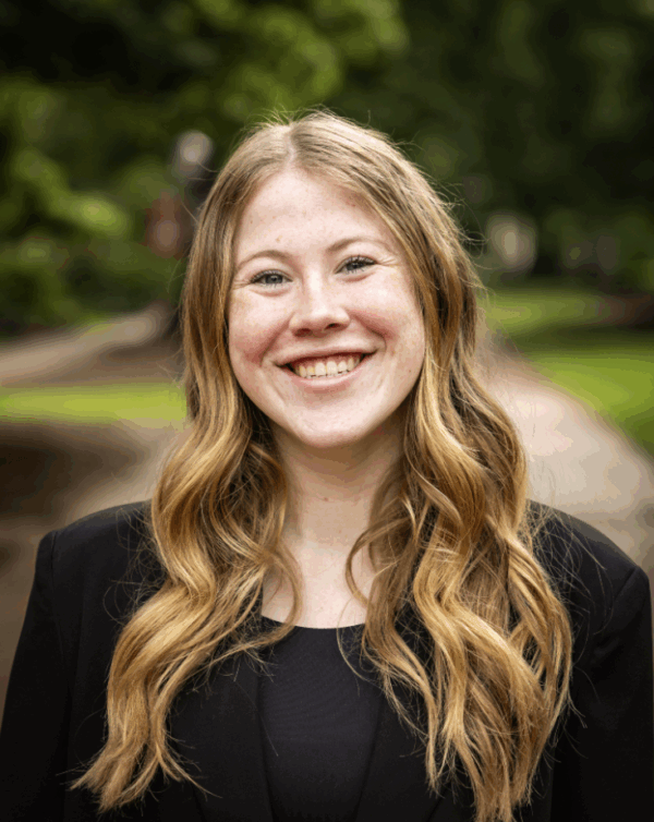 A young woman with long, wavy blonde hair smiles warmly at the camera, wearing a black blazer. She stands outdoors in a park-like setting with greenery in the background.