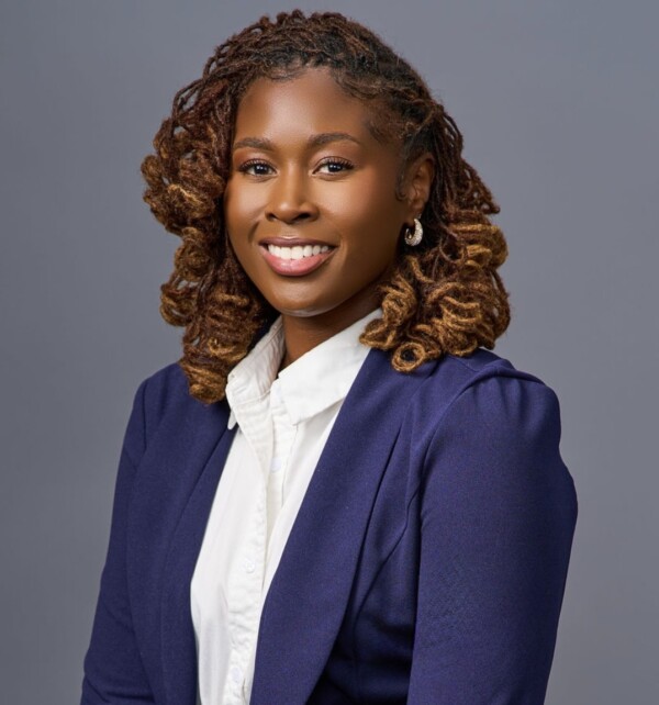 A woman with shoulder-length curly hair smiles confidently in a professional setting, wearing a navy blazer over a white blouse against a gray background.