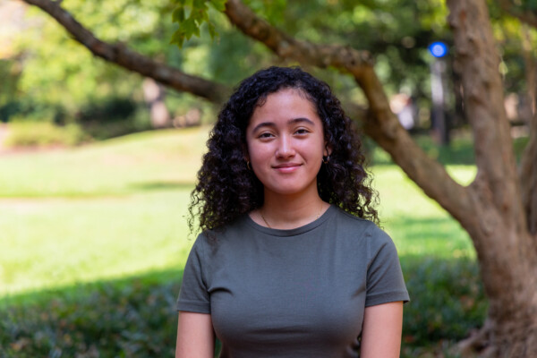 A young woman with curly hair is standing outdoors in front of a blurred green background, smiling softly. She is wearing a light green t-shirt and has small hoop earrings. A tree is partially visible behind her.