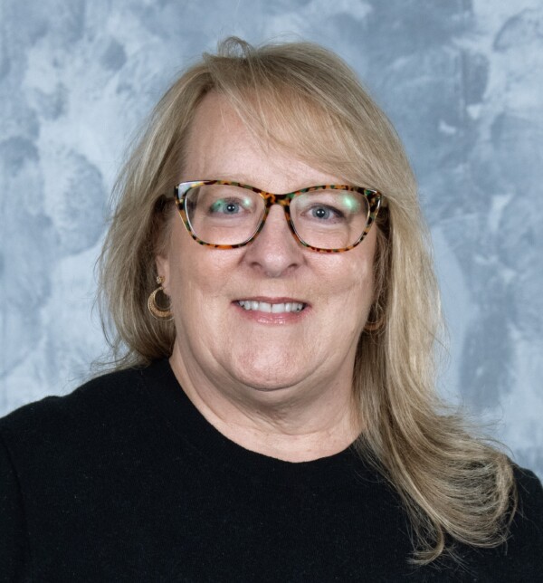 A middle-aged woman with shoulder-length blonde hair, wearing tortoiseshell glasses and gold hoop earrings, smiles at the camera against a gray textured background.