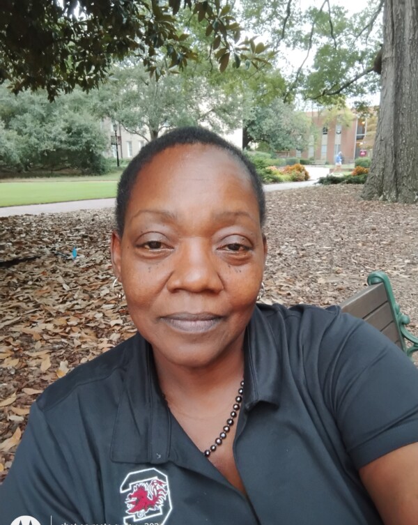 A close-up portrait of a woman sitting outdoors, wearing a black polo shirt with an emblem featuring a gamecock. She has a serious expression and is surrounded by greenery and fallen leaves in a park-like setting.