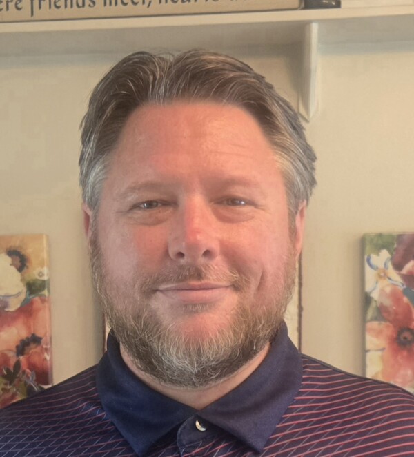 A man with a beard and short, styled hair smiles at the camera, wearing a dark polo shirt with a pattern. The background features floral wall art and a sign that reads 'Where friends meet, hearts warm.'