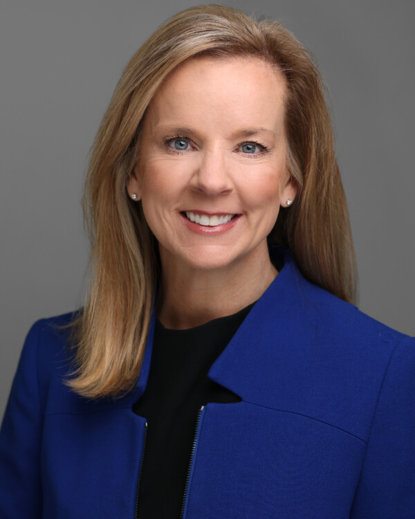 A woman with shoulder-length blonde hair, wearing a bright blue blazer over a black top, smiles warmly at the camera against a neutral gray background.