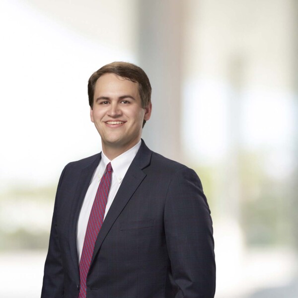 A young man in a dark suit with a white shirt and a patterned red tie is smiling confidently while standing against a softly blurred background that suggests a professional setting.