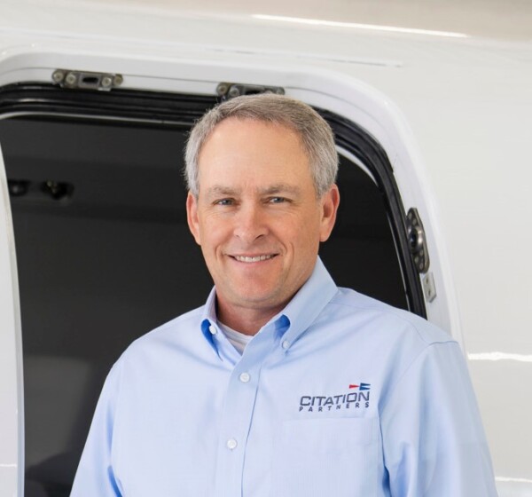 A smiling middle-aged man wearing a light blue button-up shirt with 'CITATION PARTNERS' embroidered on it, standing in front of an open aircraft door.