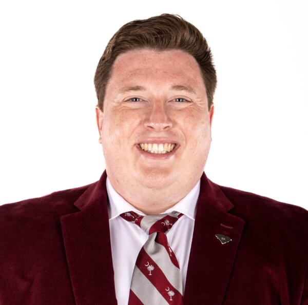 A smiling man with short, styled hair, wearing a dark burgundy blazer and a striped tie featuring a design of crescent moons and tree symbols, on a white background.