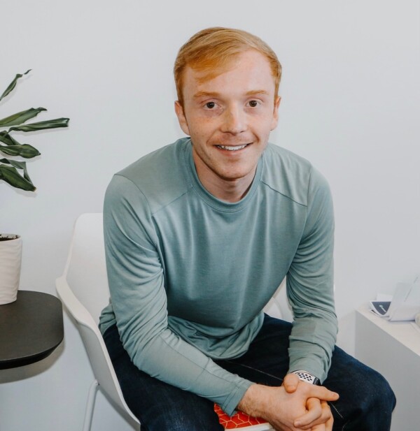 A young man with short, light brown hair smiles while sitting on a chair, wearing a long-sleeved teal shirt and dark jeans, with a potted plant and a small table visible in the background.