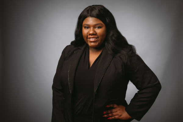 A woman with long black hair, wearing a black blazer over a black top, smiles confidently while posing with one hand on her hip against a soft gray background.