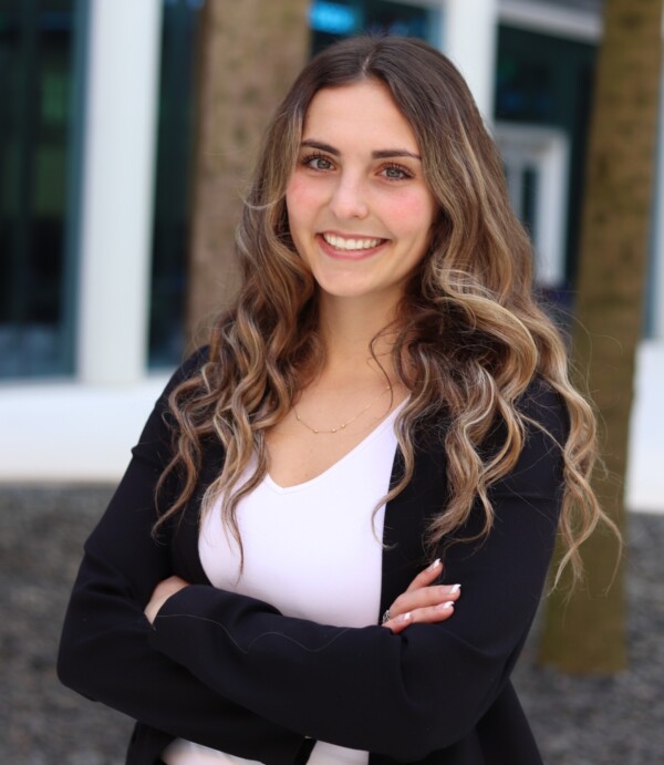 A young woman with long, wavy hair smiles confidently while crossing her arms. She is wearing a black blazer over a white top and stands in front of tall palm trees and a modern building background.