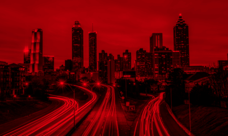 A dramatic view of a city skyline at dusk, featuring tall buildings illuminated in red. The foreground shows a highway with streaks of red light from moving vehicles, while the cityscape includes a mix of modern skyscrapers.