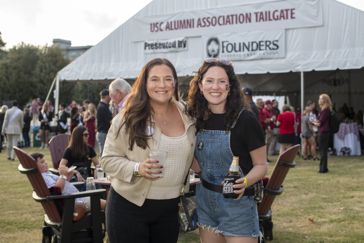 Two women pose smiling at a USC Alumni Association tailgate event, with a large white tent in the background featuring the event name. One woman wears a beige jacket and holds a drink, while the other wears a black shirt and denim overalls holding a beverage. In the background, attendees are mingling on a grassy area.
