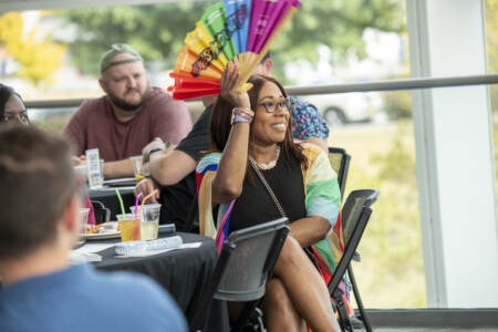 A woman with long brown hair and glasses smiles while holding a colorful fan at an outdoor event. She is wearing a black outfit and a multi-colored wrap. In the background, a group of people is seated at tables, some holding drinks.