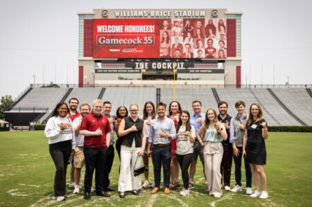 A group of 16 people stands on the field of Williams-Brice Stadium, posing for a photo in front of a large digital screen displaying "WELCOME HONOREES! Gamecock 35." The group displays a mix of casual and semi-formal clothing, with some individuals giving thumbs-up gestures. The stadium bleachers are visible in the background along with goalposts.