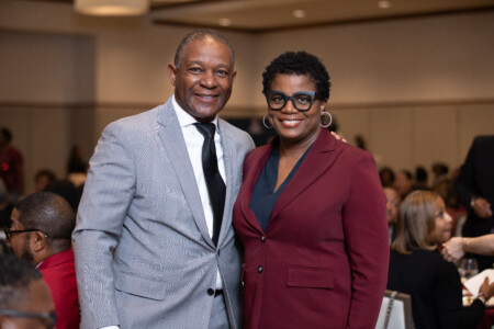 A man and a woman pose together for a photo at an event. The man wears a light gray checkered suit with a black tie, while the woman is dressed in a maroon blazer. Both are smiling, with a soft-focus background showing other attendees at tables in a banquet setting.