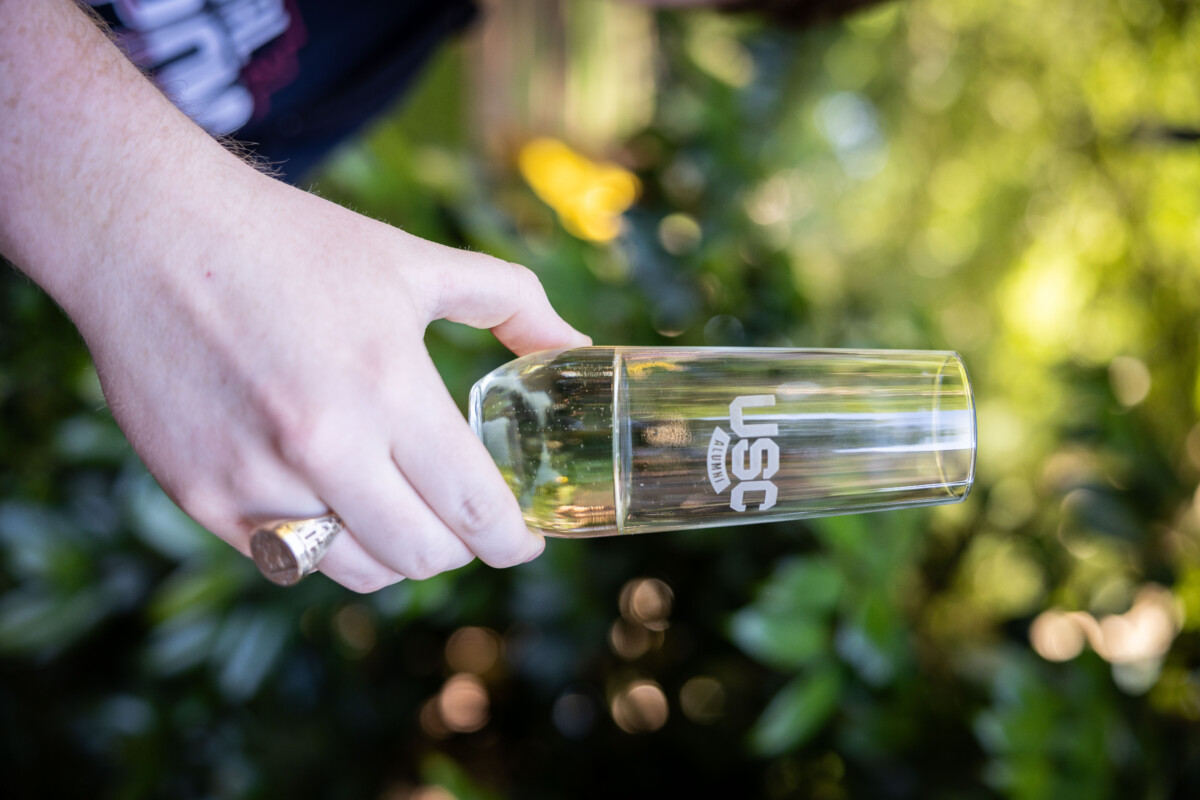 A person holding a clear glass of sparkling beverage inscribed with 'USC ALUMNI' against a blurred green outdoor background with hints of natural bokeh.